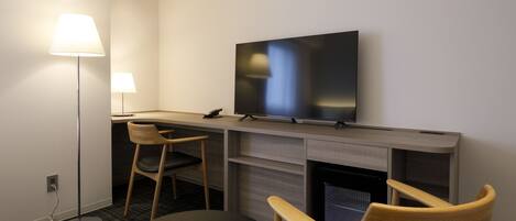 Una habitación de hotel moderna con una televisión de pantalla plana montada en la pared, un escritorio con una silla, una lámpara de pie y una mesa redonda.