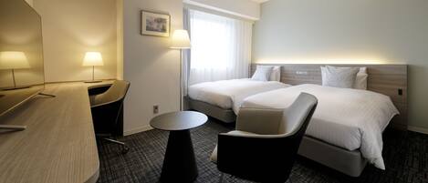 A hotel room with two beds, a desk, a chair, a small table, and a window with curtains.
