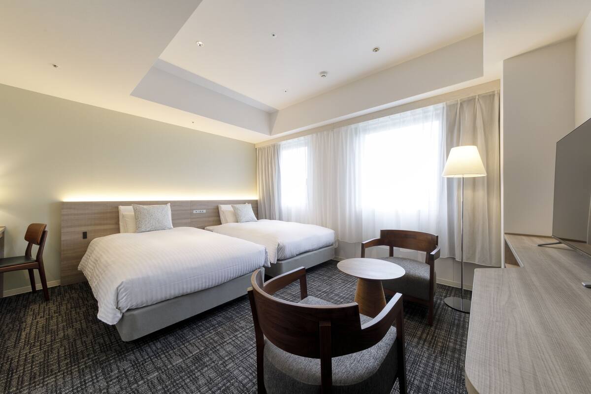 A hotel room with two beds, a desk, a chair, a small table, and a TV.