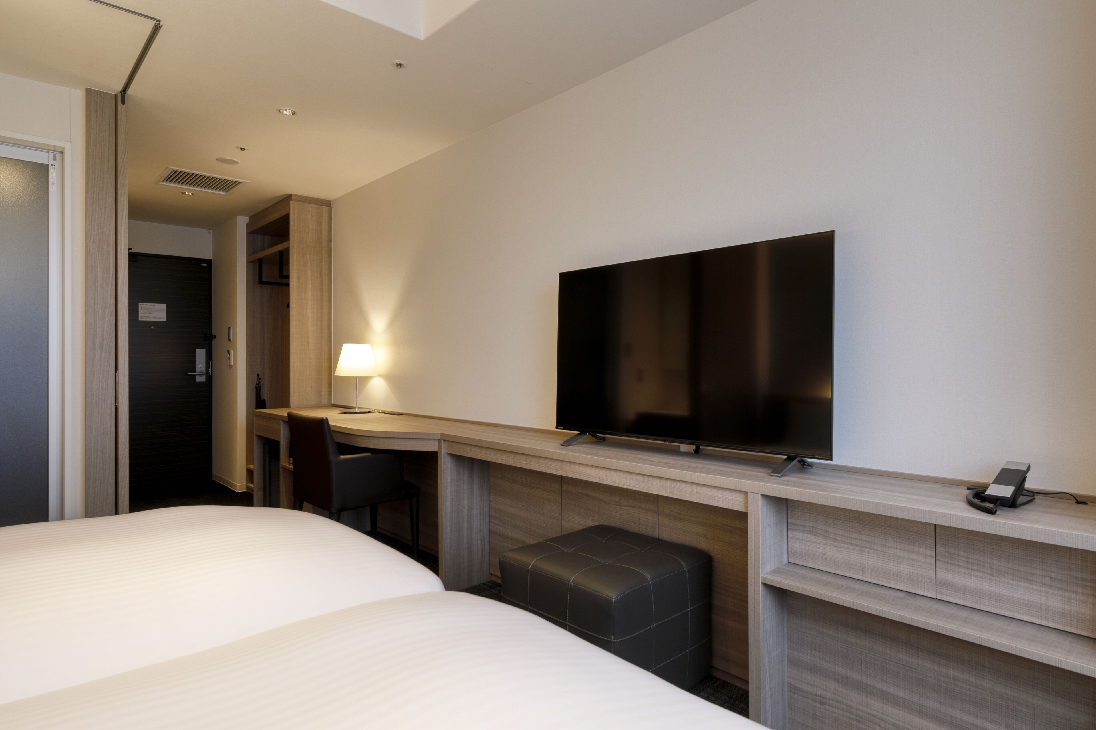 A hotel room with a bed, a desk with a chair, a television mounted on the wall, and a small ottoman.