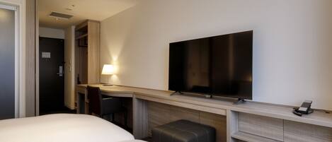 A hotel room with a bed, a desk with a chair, a television mounted on the wall, and a small ottoman.