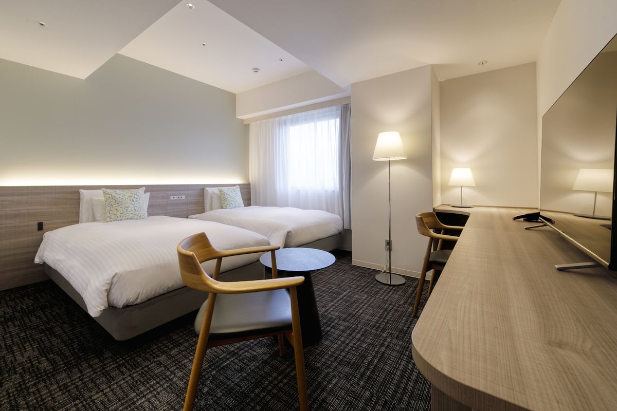 A hotel room with a bed, two chairs, a small table, a desk, a lamp, and a mirror.