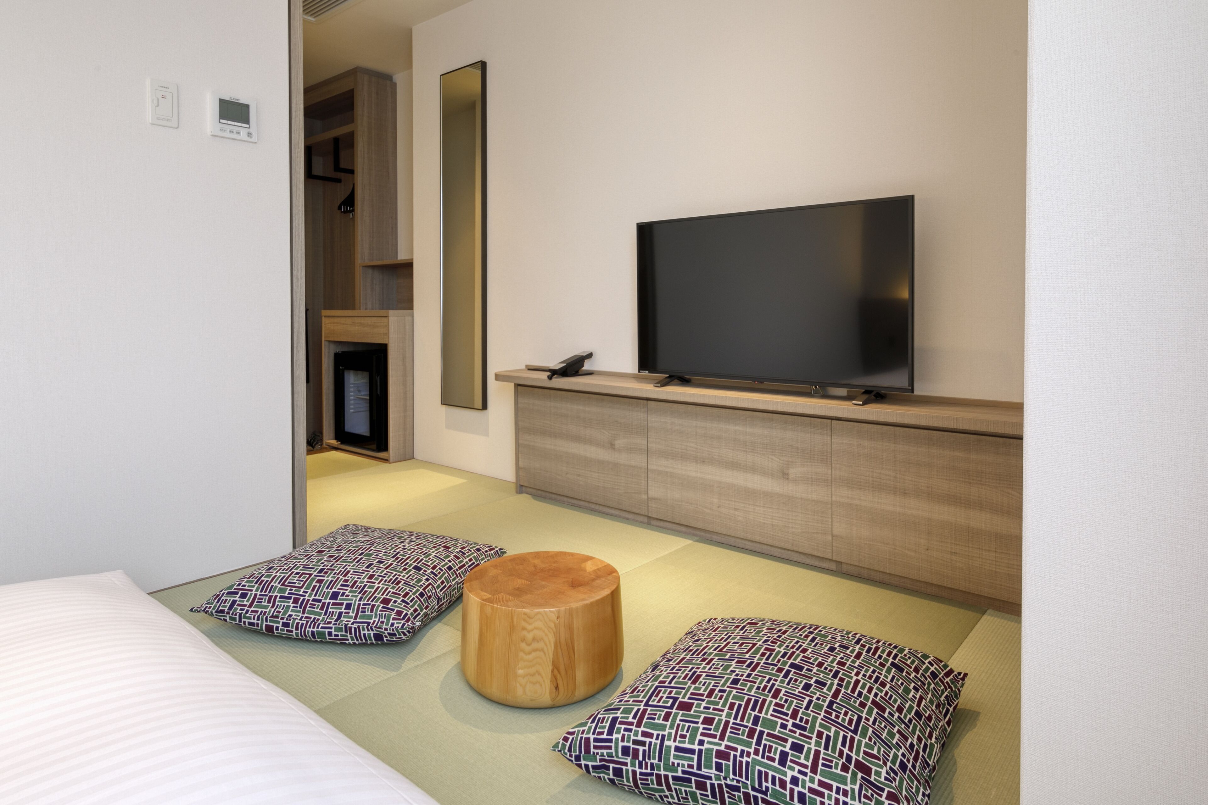A modern hotel room with a large flat-screen TV mounted on a wooden cabinet, a bed with patterned pillows, and a small wooden stool.
