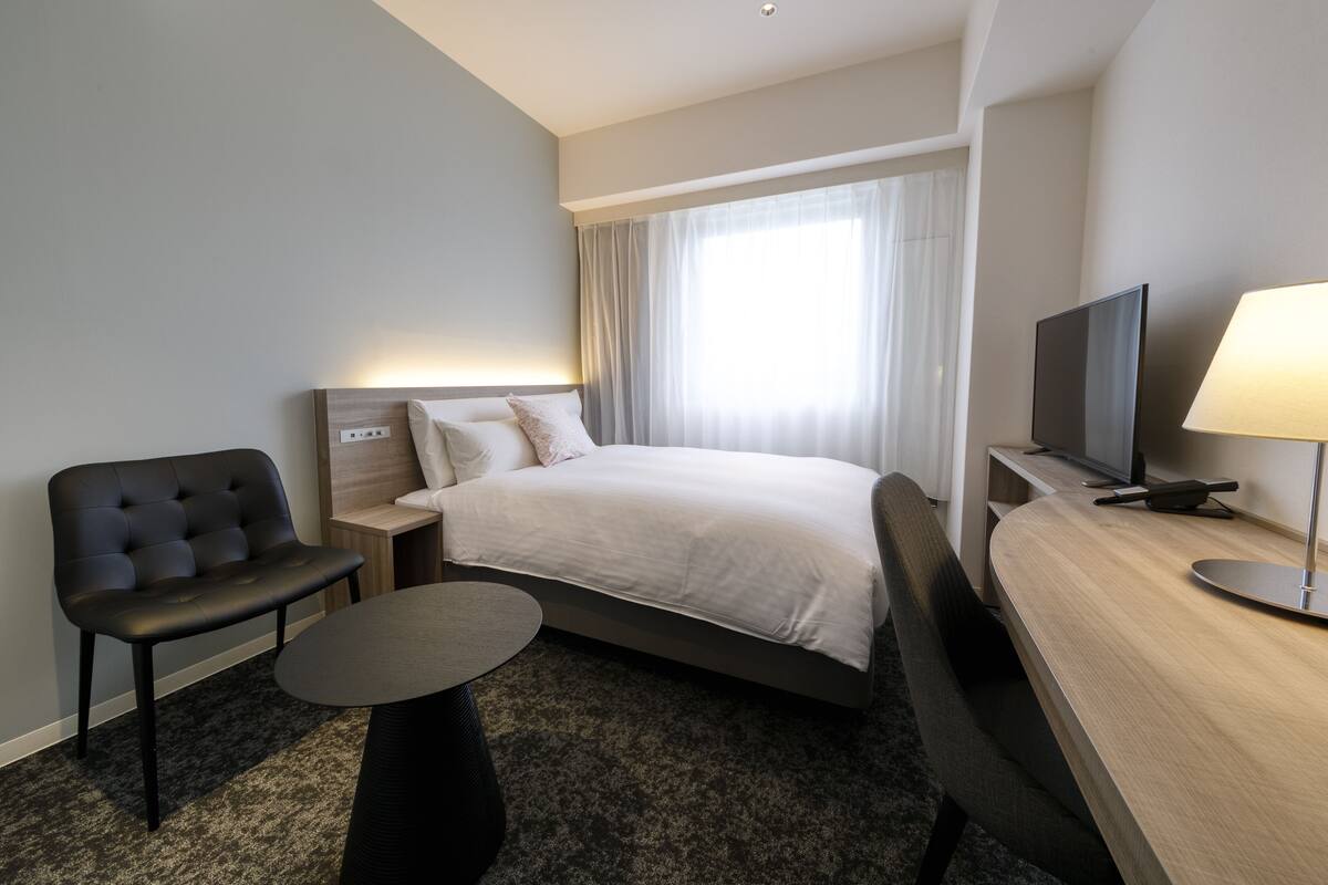 A hotel room with a bed, a desk, a chair, a TV, and a lamp.