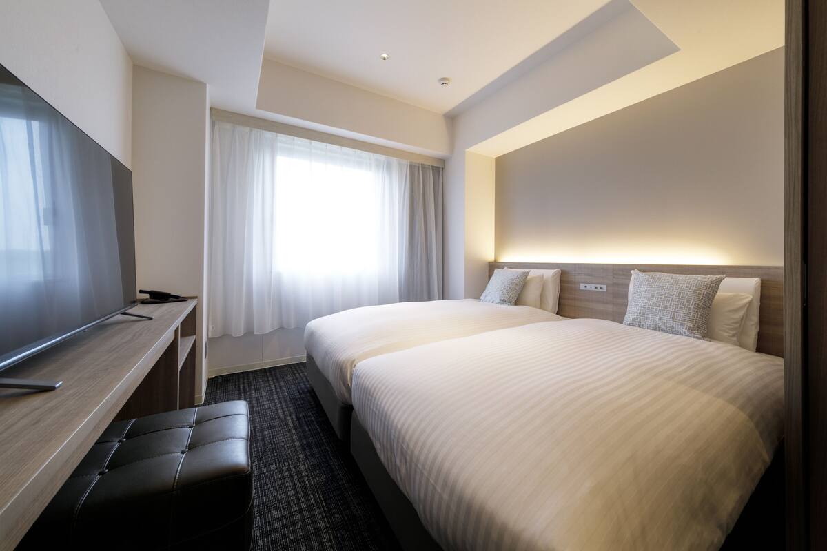 A hotel room with a large bed, a desk with a chair, a flat-screen TV, and a window with curtains.