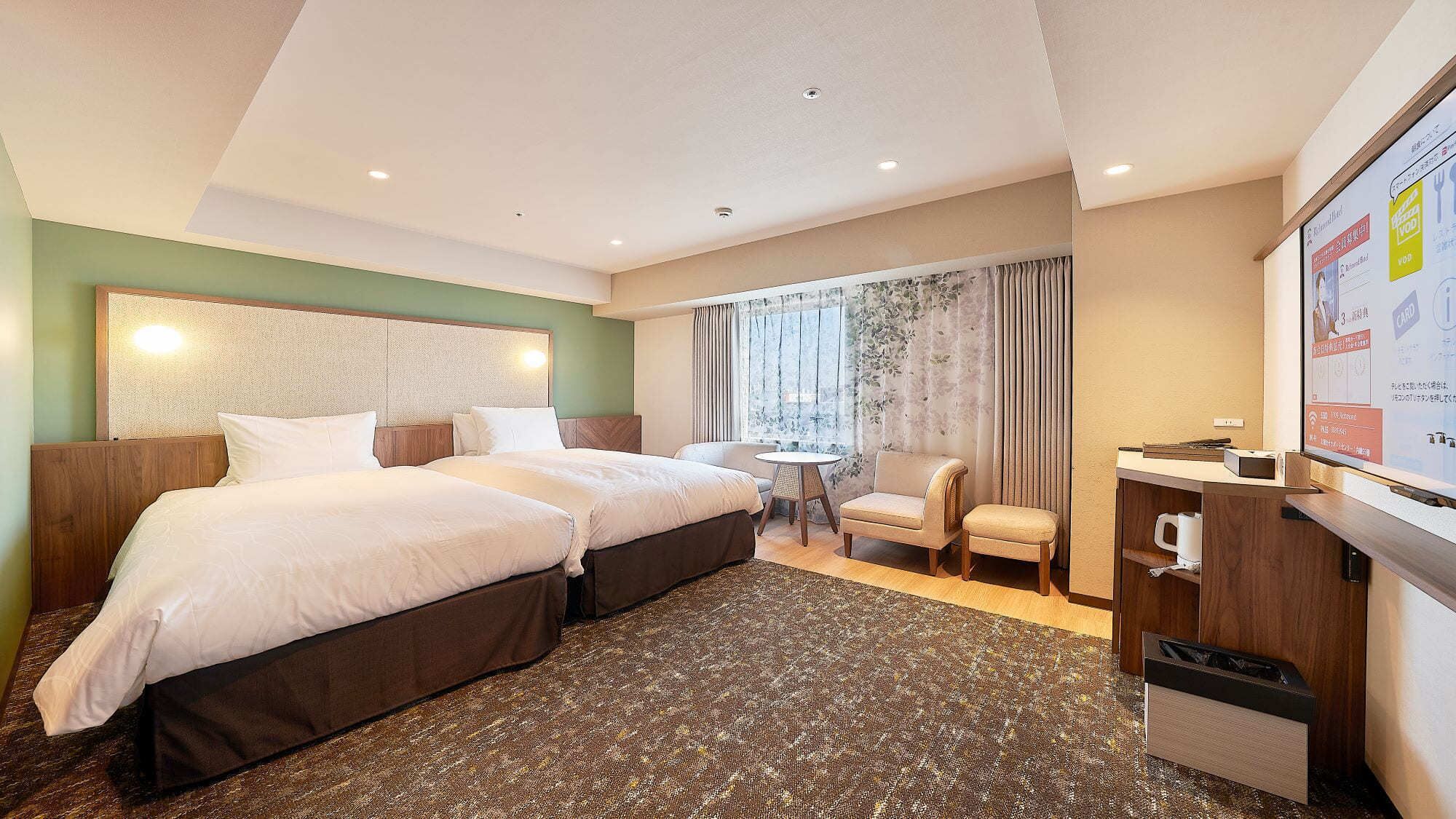 Quarto de hotel com duas camas, uma mesa pequena, uma cadeira, uma escrivaninha com um computador e vista para a cidade pela janela.
