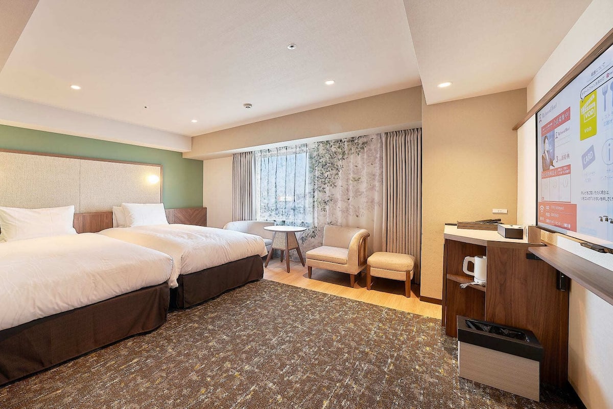 Een hotelkamer met twee bedden, een kleintje tafeltje, een stoel, een bureau met een computer en uitzicht op de stad door het raam.