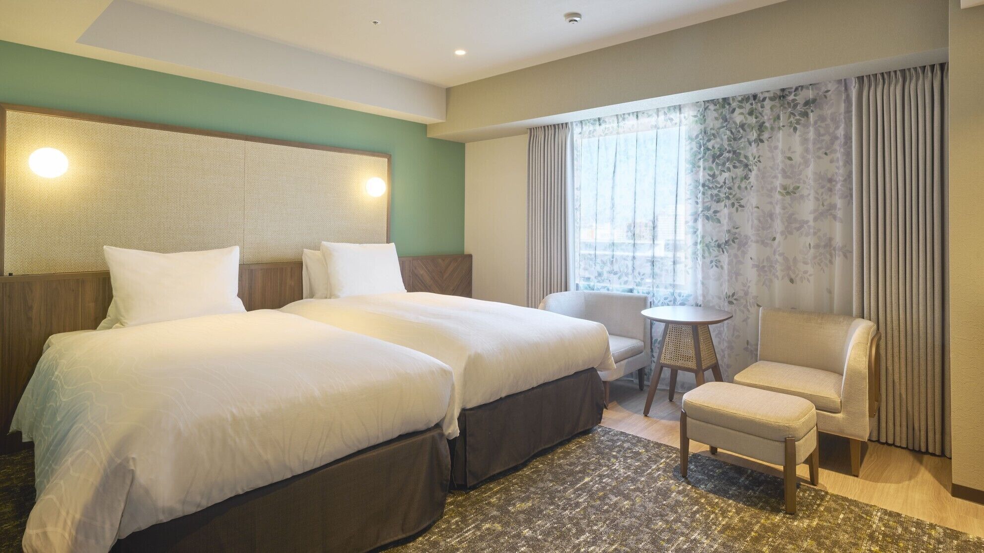 Quarto de hotel com duas camas, uma mesa redonda pequena, uma cadeira e uma janela grande com cortinas.
