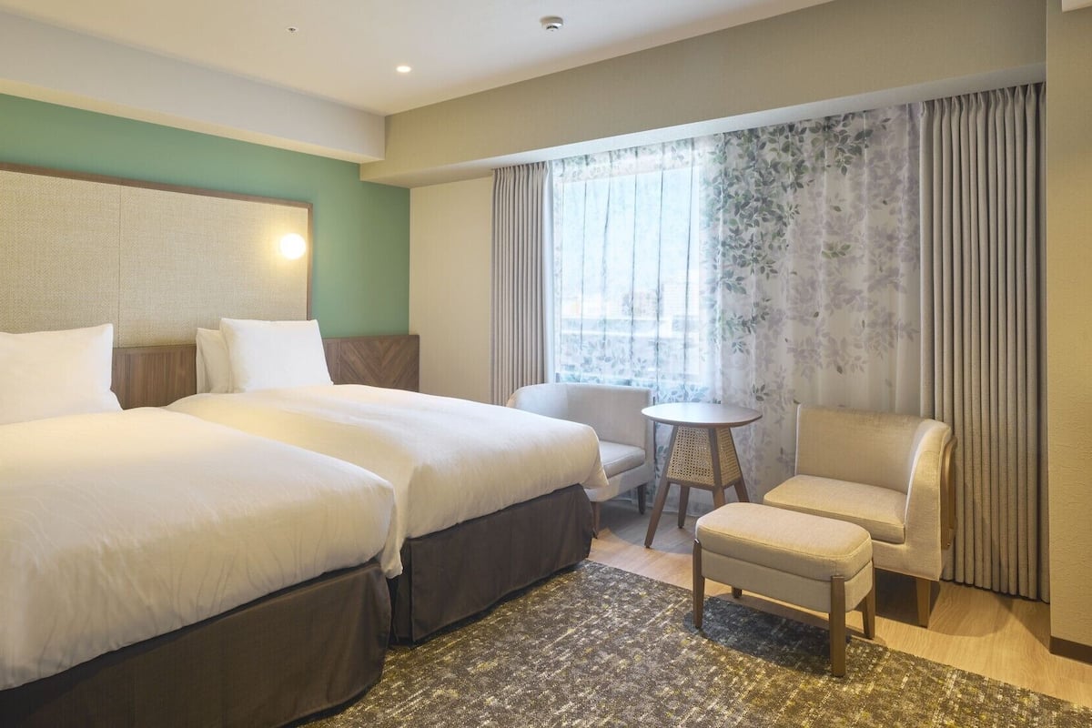 Een hotelkamer met twee bedden, een klein rond tafeltje, een stoel en een groot raam met gordijnen.