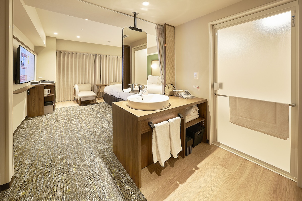 A modern hotel room with a wooden vanity, a large mirror, a flat-screen TV, a comfortable chair, and a large bed.