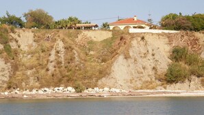 A coastal house with a red-tiled roof, situated on a hillside with a rocky shoreline in the foreground. - SeaFront Stone Villa with private access to the Beach (Άγιος Σώστης)