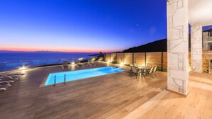 A rooftop pool area with a wooden deck, lounge chairs, and a view of the ocean at dusk. - SeaFront Stone Villa with private access to the Beach (Άγιος Σώστης)