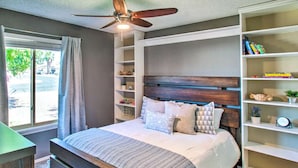 Ein Schlafzimmer mit einem Bett, einem Deckenventilator, einem Fenster mit Vorhängen und eingebauten Regalen mit Büchern und Dekoration.