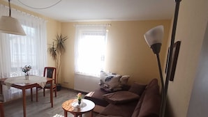 Een woonkamer met een bank, een salontafel, een eettafel met stoelen, en een groot raam met gordijnen.