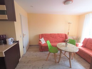 A living room with a red sofa, a green chair, a round table, and a white kettle on a shelf.