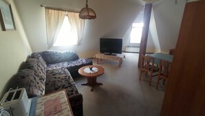 Een woonkamer met een bank, een klein rond tafeltje, een televisie en een eethoek met een tafel en stoelen.