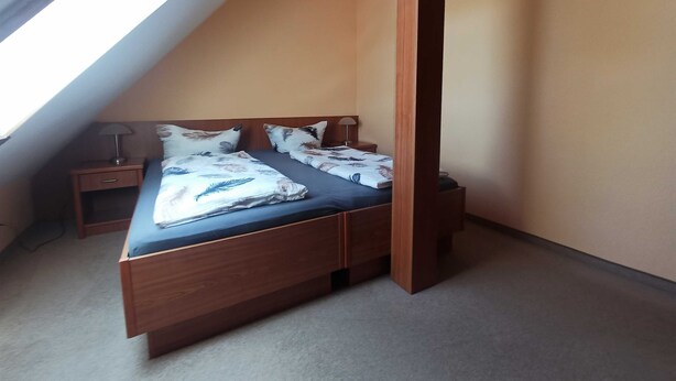 Een kleine slaapkamer met schuin dak, een houten bed, nachtkastje en een lamp.