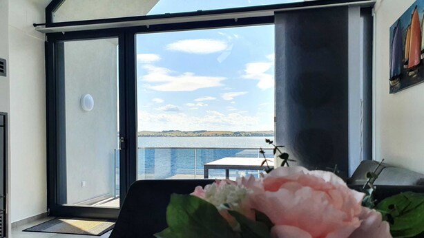 Una habitación con un gran ventanal corredero que da a un balcón con vistas al agua y las colinas.