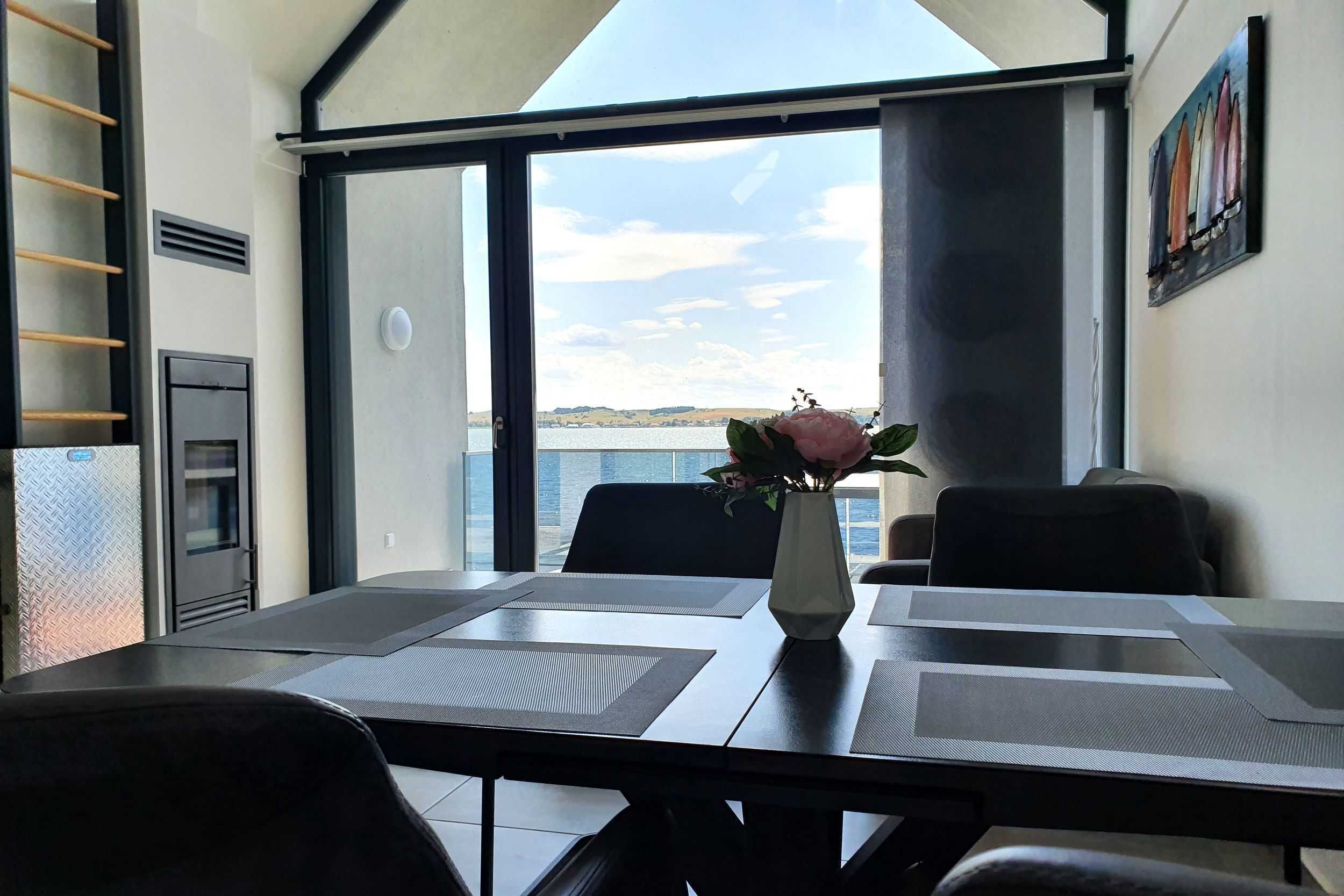 A dining area with a table set for four, a vase with flowers, and a view of the sea through large windows.