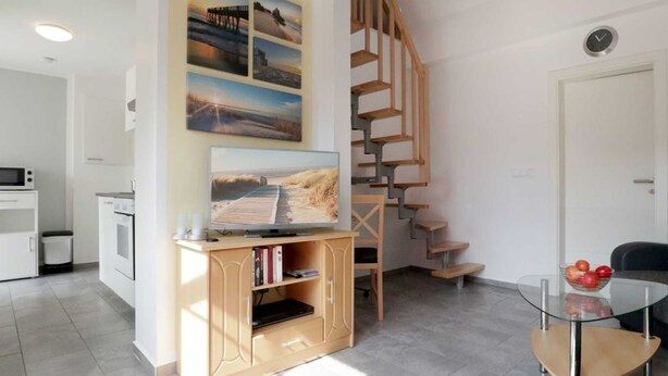 A modern living room with a wooden staircase, a flat-screen TV on a wooden console, a glass coffee table, and a view of the outside. - Troja Ferienwohnung, Deutschland (Kalkhorst)