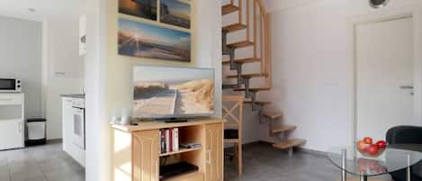 A modern living room with a wooden staircase, a flat-screen TV on a wooden console, a glass coffee table, and a view of the outside.
