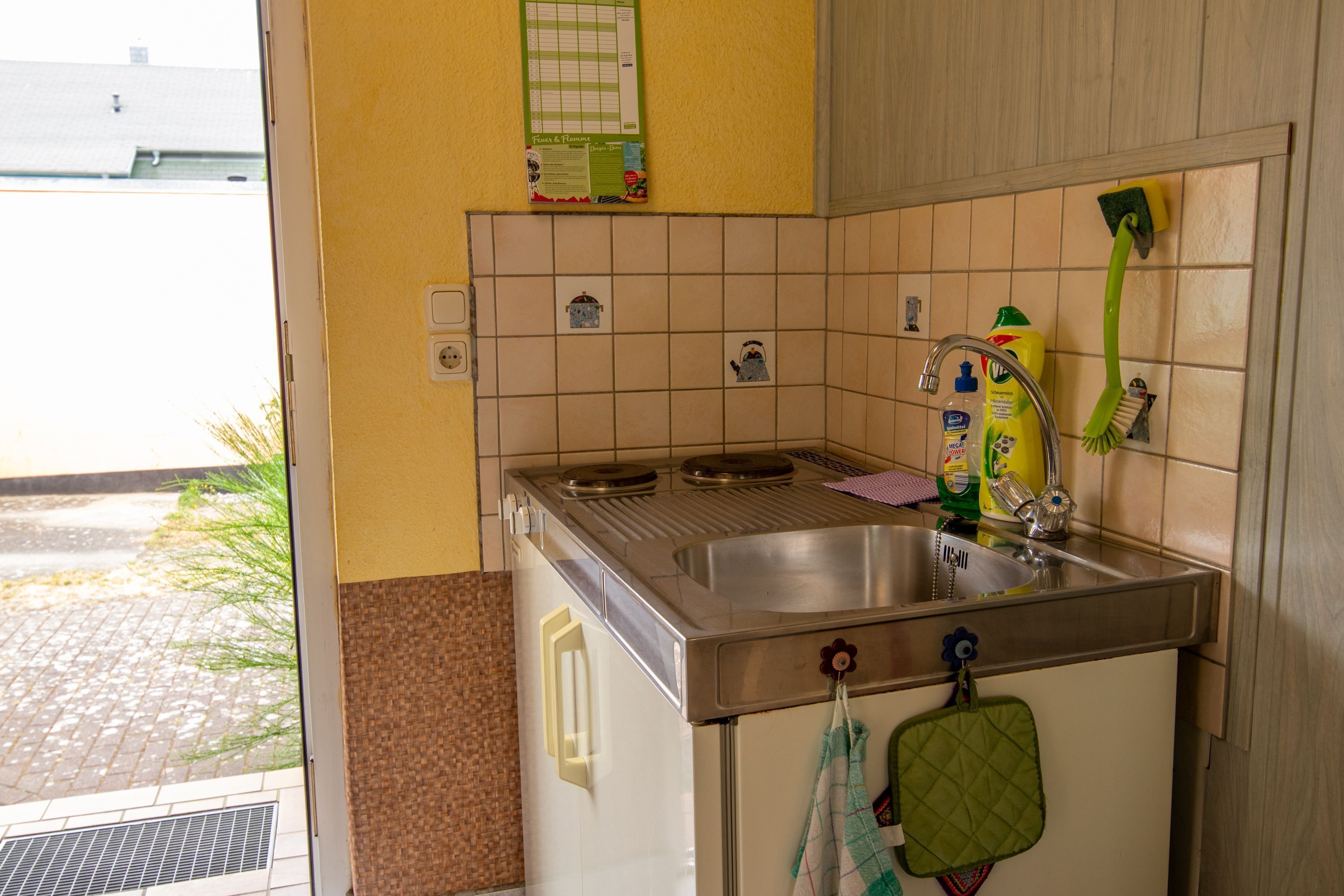 A kitchen with a stainless steel sink, a green-handled brush, and a tiled backsplash.