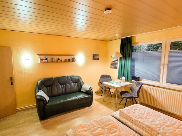 Ein Zimmer mit Holzboden, einem schwarzen Ledersofa, einem kleinen Tisch mit zwei Stühlen, einem Bett mit rosa Matratze und einem Fenster mit Jalousien.