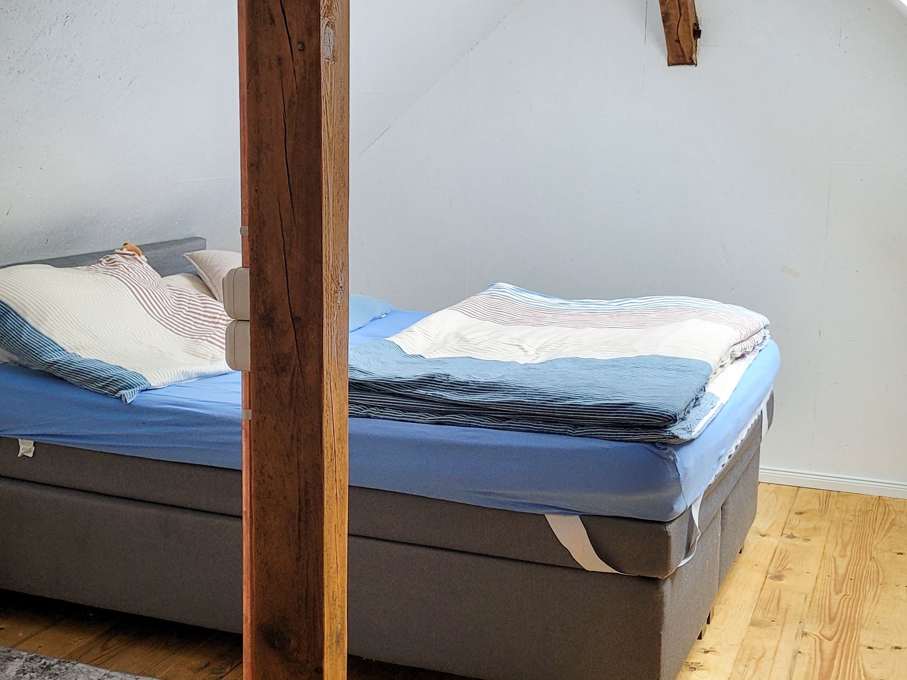 A bed with a grey mattress and blue bedding, a wooden headboard, and a plain wall.