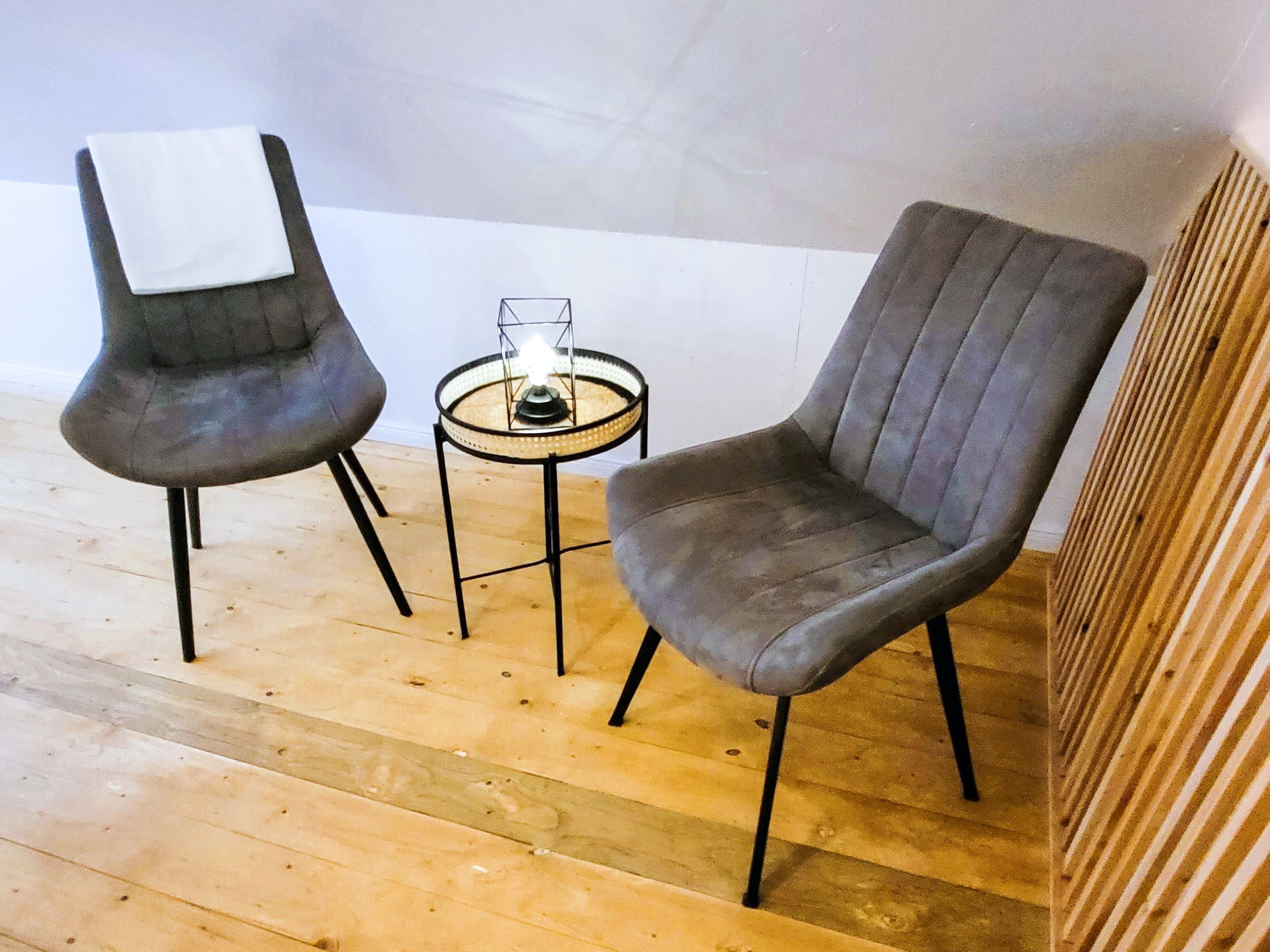 Dos sillones grises tapizados con patas de metal negro, una mesita redonda con superficie de vidrio y una vela encendida en el centro.