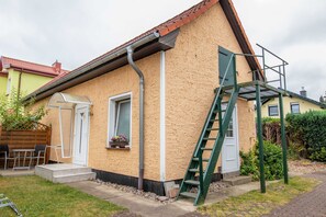 Una casa con un pequeño jardín delantero, cerca de madera, una escalera de metal y una ventana con un balcón de flores.