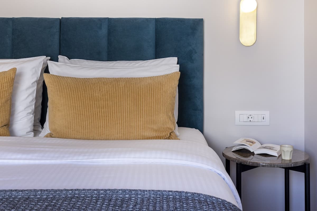 Ein ordentlich bezogenes Bett mit blauem Kopfteil, weißen und senffarbenen Kissen und einer gemusterten Tagesdecke. Ein kleiner Nachttisch mit Buch und Tasse ist sichtbar.