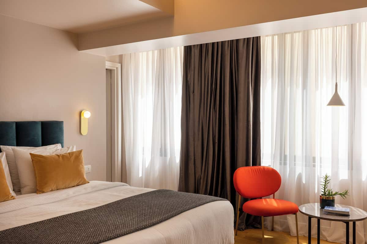 Ein Hotelzimmer mit einem Bett, einem roten Sessel, einem kleinen Tisch mit Pflanze und einem Fenster mit Vorhängen.