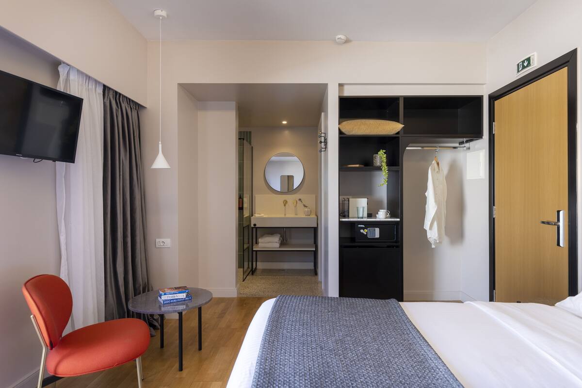 Ein Hotelzimmer mit einem Bett, einem Fernseher, einem roten Sessel, einem kleinen Tisch, einem Spiegel, einem Kleiderschrank und einem durchgegangenen Badezimmer.