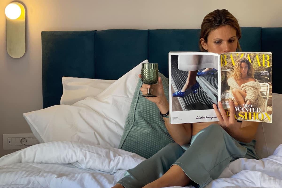 Eine Person liegt auf einem Bett, neben ihr ein Magazin, ein Glas Wasser und eine Wandleuchte.