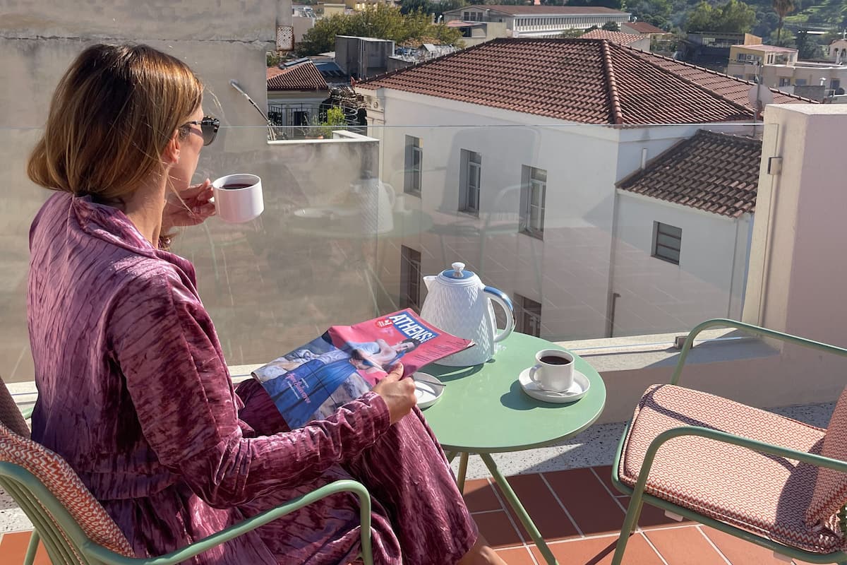 Eine Person sitzt auf einem Balkon und blickt auf eine historische Stadtansicht, darunter ein markanter Hügel mit antiken Ruinen.