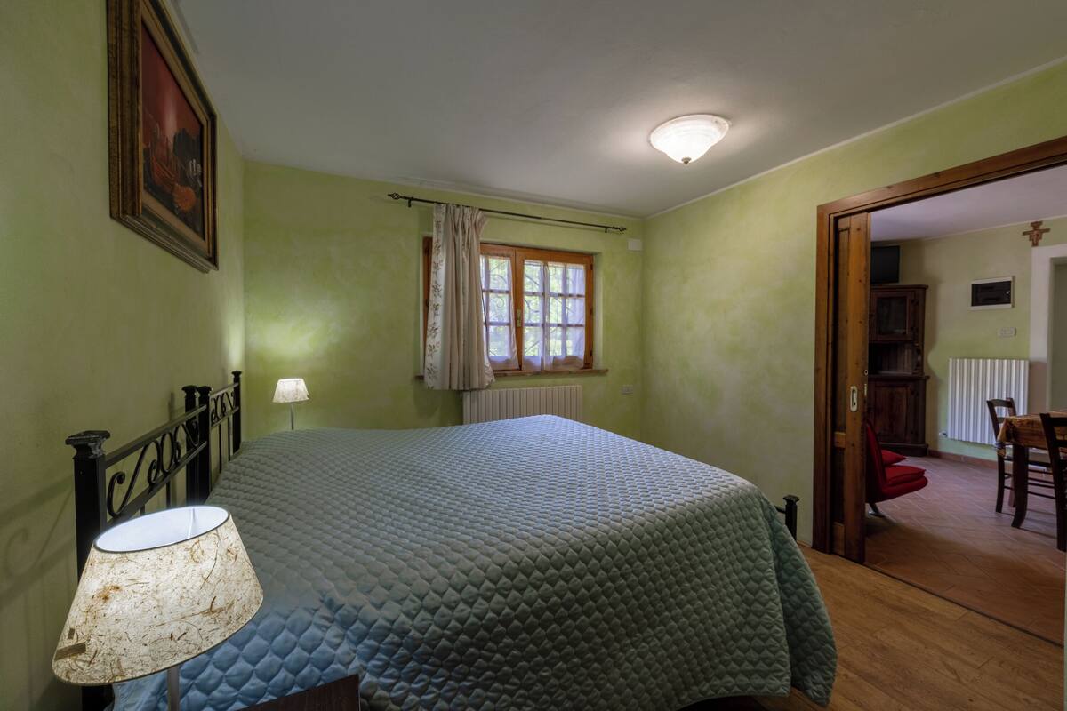 A bedroom with a bed, a lamp, a window with curtains, and a door leading to another room.