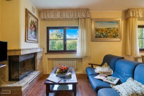 A living room with a fireplace, a sofa, a coffee table with a fruit arrangement, and two windows with curtains.