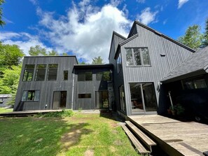 A modern two-story house with a flat roof, large windows, and a wooden deck.