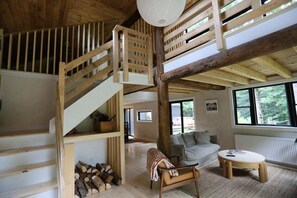 Un interior de madera con una escalera, un sofá, una mesa redonda y un ventanal con vistas a árboles.