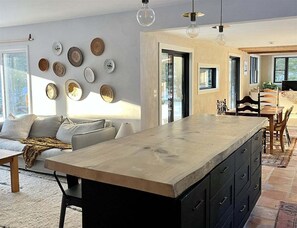 A modern kitchen with a large island, a dining area with a table and chairs, and a living area with a sofa and decorative plates on the wall.