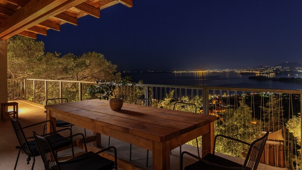A wooden table with chairs, a railing, and a view of a cityscape and water at night. - Greenstone Villas (Corfu)