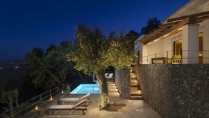 A modern outdoor pool area with a stone wall, wooden deck, and lounge chairs. - Greenstone Villas (Corfu)