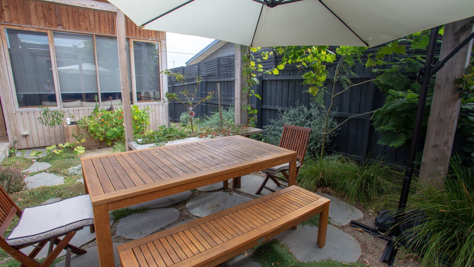 A wooden patio with a table and chairs, surrounded by a garden with various plants and a wooden fence.