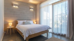 A bedroom with a wooden bed, two bedside lamps, a brick wall, and a large window with sheer curtains.