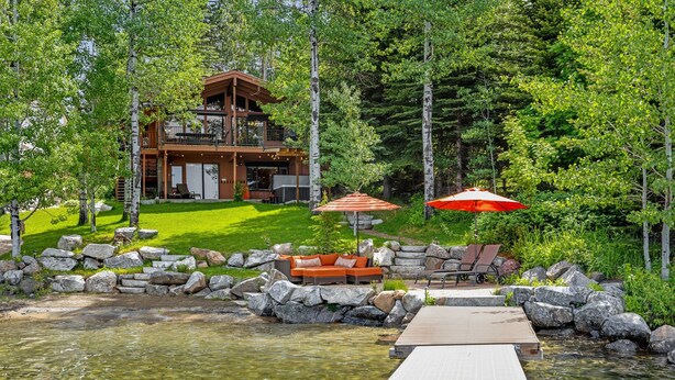 Una cabaña junto al lago con terraza, mobiliario exterior y vistas despejadas del bosque circundante.