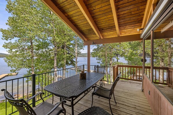 A covered outdoor deck with a table and chairs, overlooking a lake with trees and a dock. - McCall Lakefront Oasis - Family Summer Paradise - Hot tub - Dock - 4 Bedrooms (McCall)