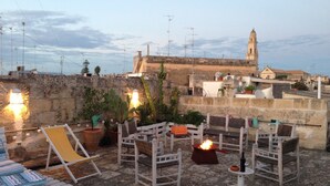 En tagterrasse med udendørs møbler, et bålsted og udsigt til en historisk bygning med et kirketårn.
