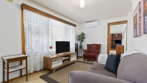 Un salon avec un téléviseur, un fauteuil rouge, un canapé, une table d’appoint en bois et une plante en pot.