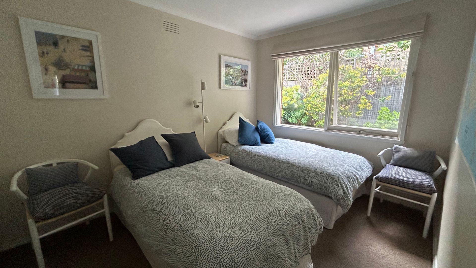 Une chambre avec deux lits simples, une chaise, une fenêtre donnant sur un espace verdoyant et deux tableaux encadrés accrochés au mur.
