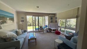 A spacious living room with a large window, a sofa, a coffee table, and a dining area with a striped tablecloth. - Dune Retreat - Point Lonsdale (POINT LONSDALE)